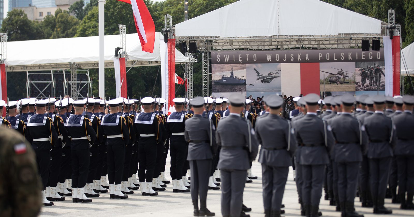 Kompanie honorowe Wojska Polskiego w szyku przed sceną z napisem Święto Wojska Polskiego, w tle polska flaga i wieżowce Warszawy